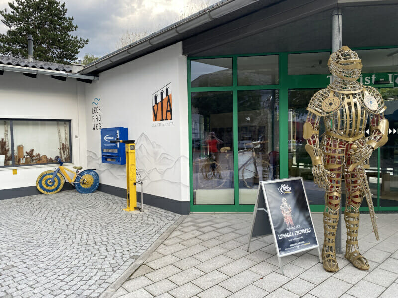 Vor den Logos des Lechradwegs & des Via Claudia Augusta Radwegs steht eine Figur mit Ritterrüstung und ein besticktes Fahrrad.
